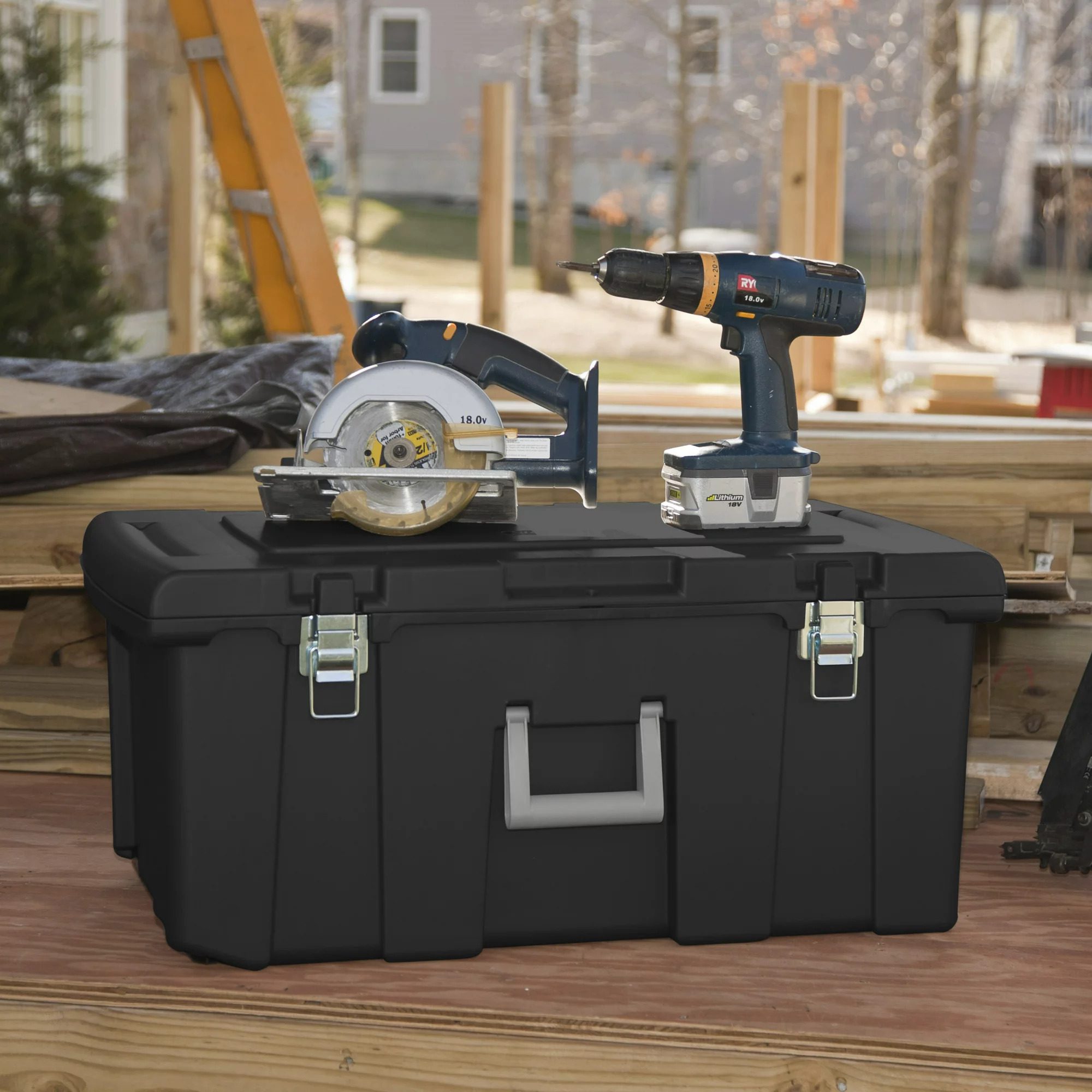 A black tool box holds a circular saw and a drill. They sit on a wooden surface with construction materials and a house in the background.
