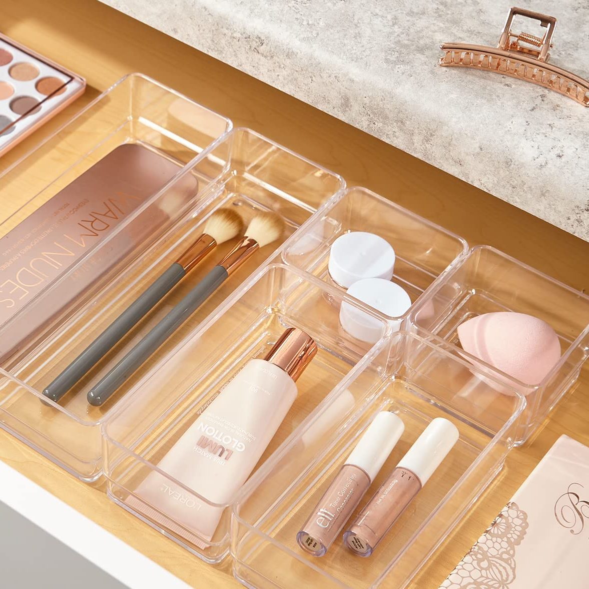 A clear organizer holds makeup brushes, sponges, and products on a light wooden surface, suggesting a tidy beauty routine in a well-lit space.