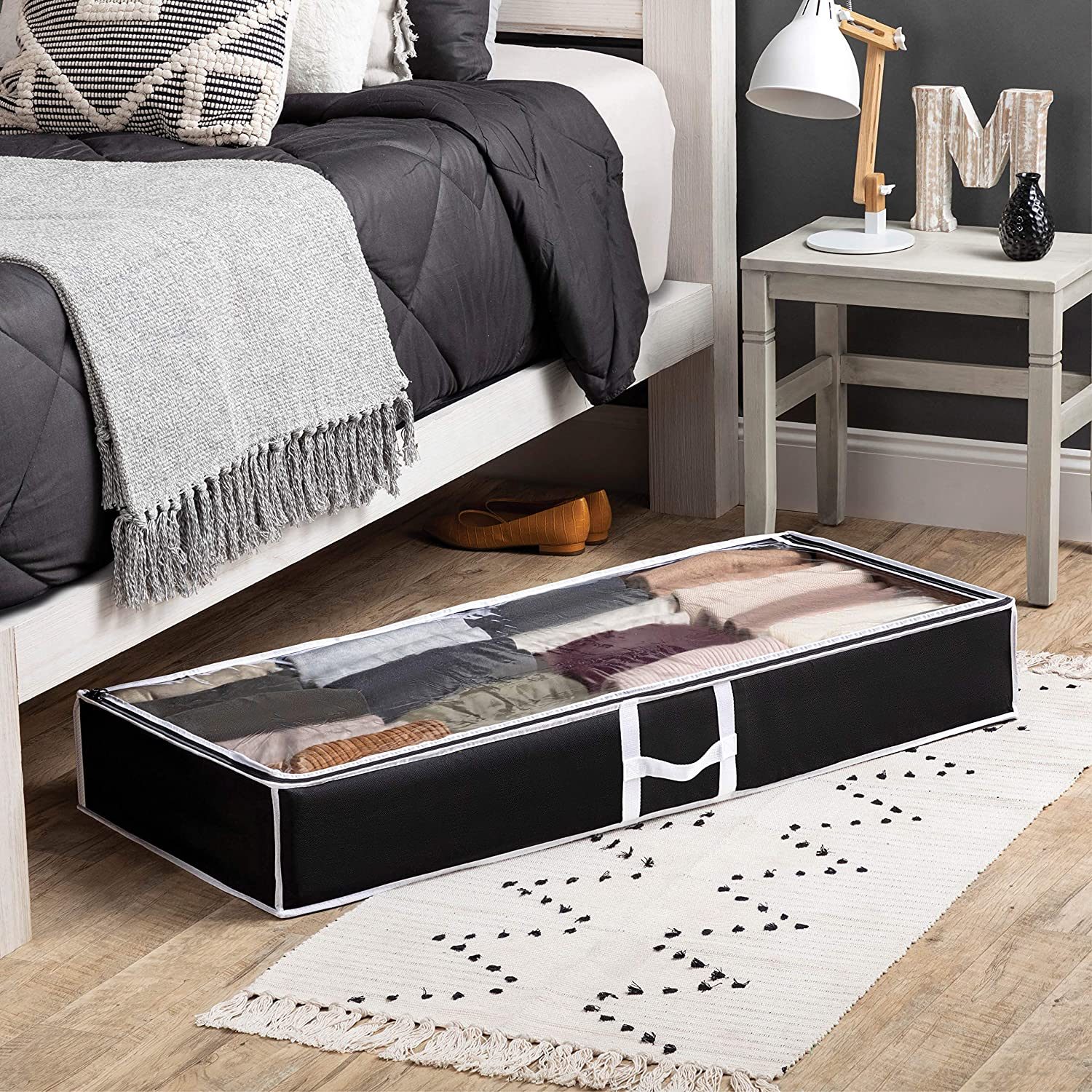 A storage container, filled with neatly folded clothes, is positioned under a bed in a stylish room featuring a rug, lamp, and decorative items.