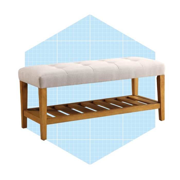 A tufted bench with a cushioned top rests on a wooden frame, featuring an open slat shelf underneath, set against a light blue geometric background.