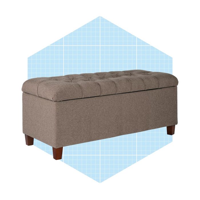A rectangular, upholstered storage ottoman rests on wooden legs, featuring a tufted top and set against a light blue grid-patterned background.