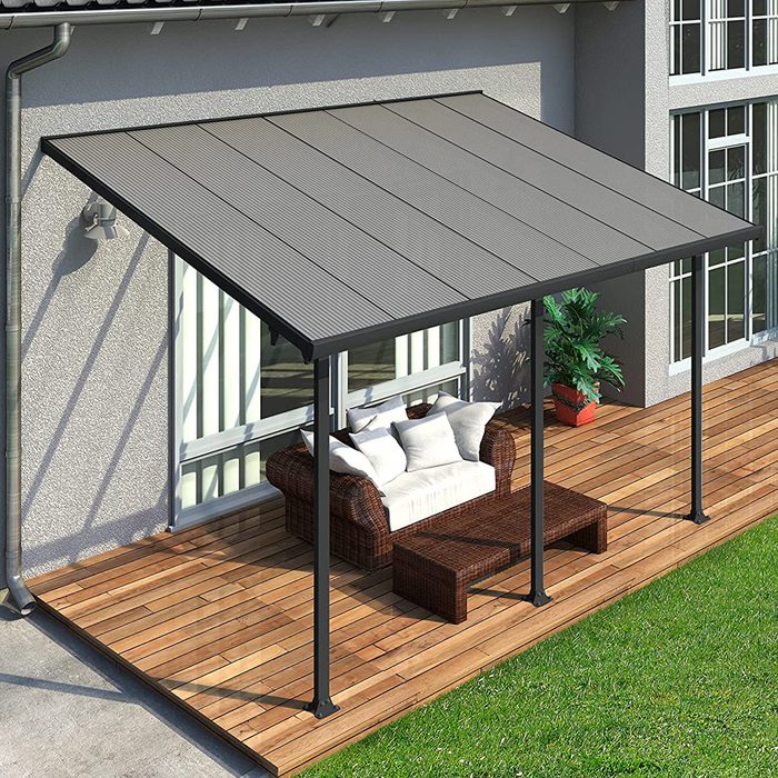 A shaded patio features a wicker seating area with cushions and a coffee table on a wooden deck, adjacent to a house with large windows.