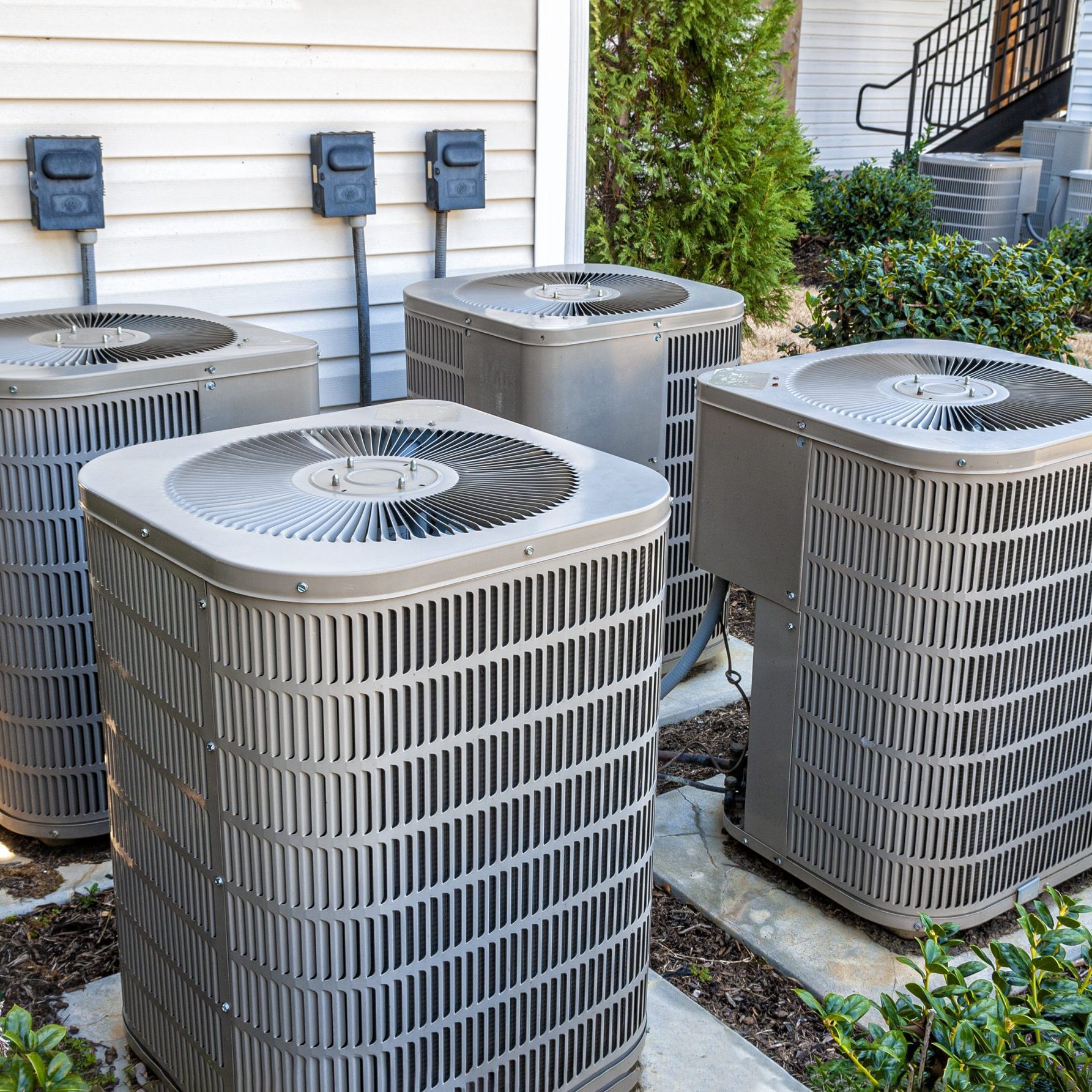 Four air conditioning units sit outside a building, connected to electrical outlets, on a stone path surrounded by low shrubs and greenery.