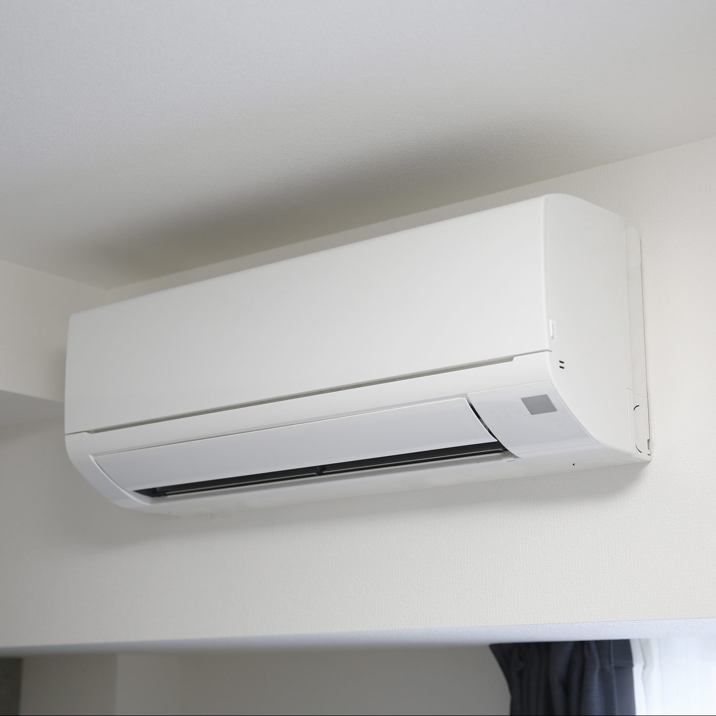 A white air conditioner mounted on a wall blows air into a modern room, which has a neutral color scheme and a window covered by dark curtains.