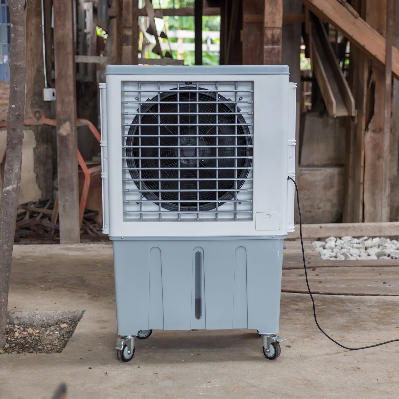 A portable air cooler is positioned on wheels, blowing air, in a partially constructed wooden building with exposed materials and scattered stones on the ground.