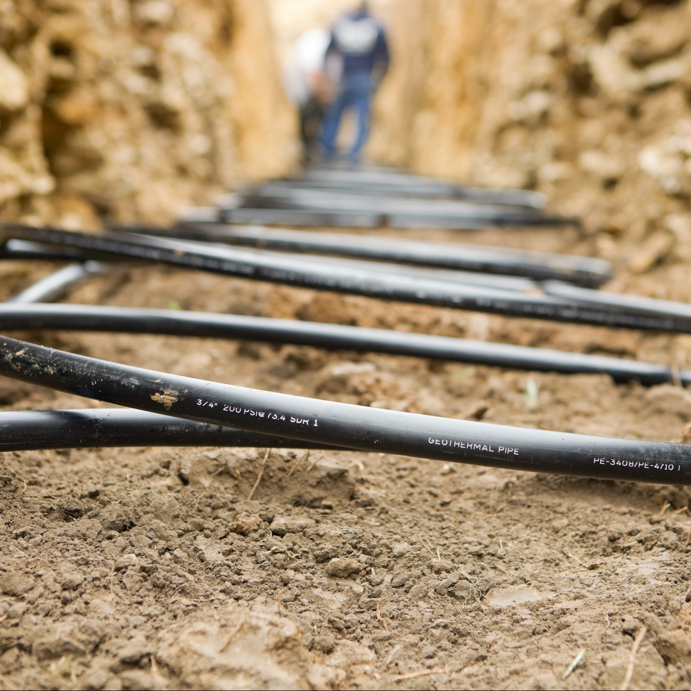Black geothermal pipes lie on disturbed soil while workers are positioned in the background, indicating installation or maintenance in a trench environment.