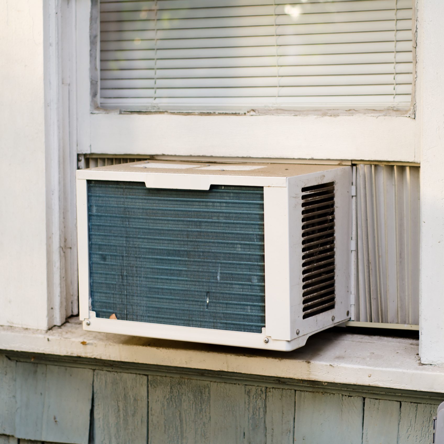An air conditioning unit is positioned in a window, drawing in outside air while surrounded by a wooden frame and nearby property exterior.