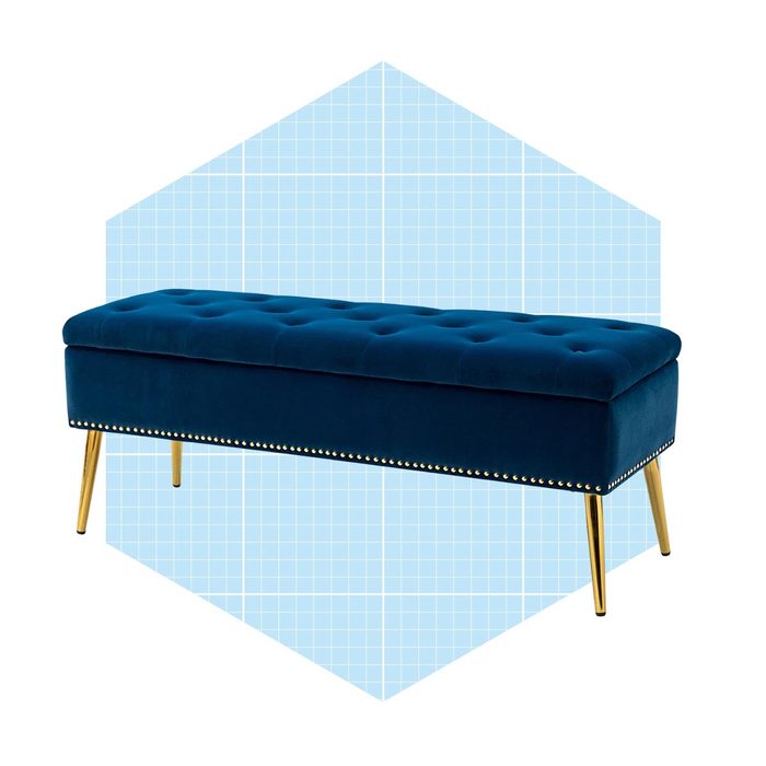 A blue velvet bench with golden legs rests against a light blue geometric background, featuring tufted upholstery and decorative nailhead trim.
