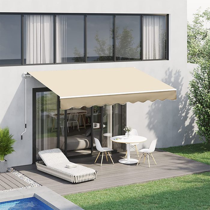 A beige awning extends over a patio with a round table and chairs, adjacent to a lounging chair by a pool, framed by green grass.