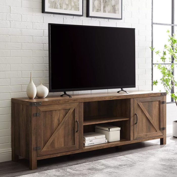 A wooden TV stand holds a large, black screen, with decorative vases beside it, set against a bright, airy room with green plants and framed artwork.