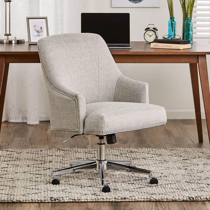 A stylish gray office chair with armrests sits on a textured rug beneath a wooden desk, surrounded by a laptop, clock, and decorative items.
