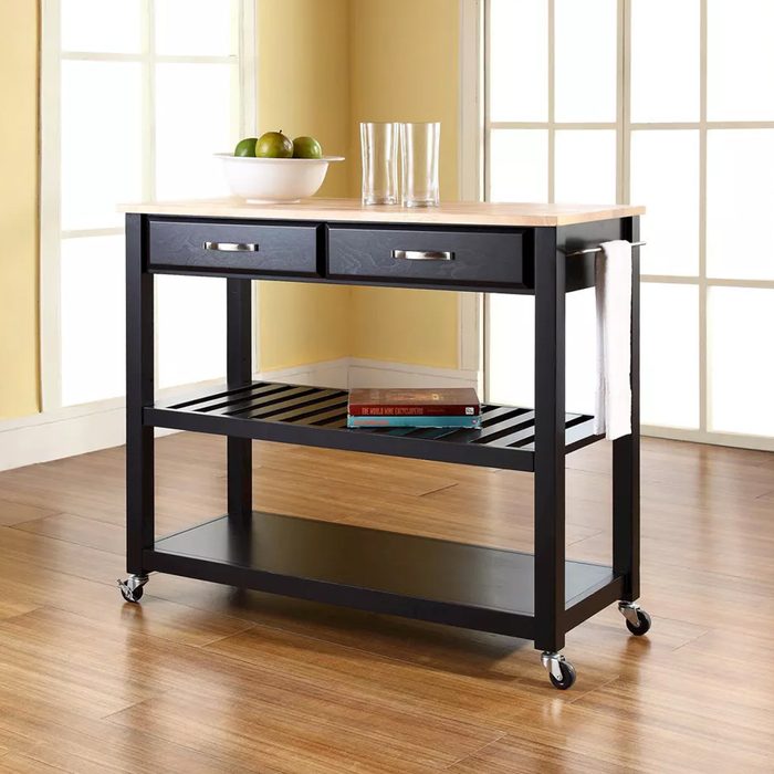 A kitchen cart with a wooden top and two drawers is positioned indoors, featuring a bowl of fruit, glasses, and books on a lower shelf.