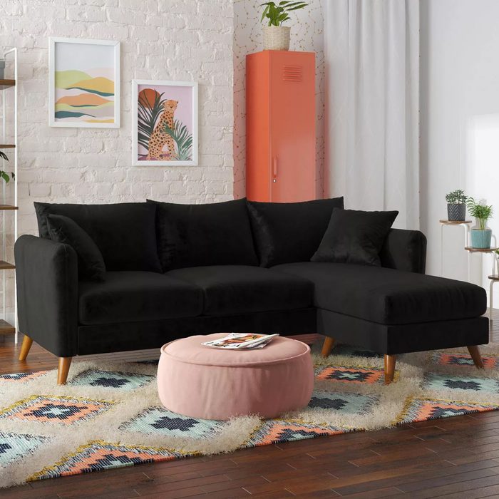 A black sectional sofa sits on a colorful patterned rug, accompanied by a pink ottoman. The backdrop features white brick walls and decorative elements.