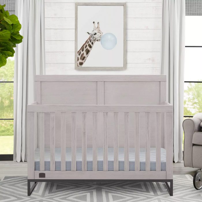 A light-colored crib stands in a bright room, with a giraffe painting blowing a bubble above it and a patterned rug beneath.