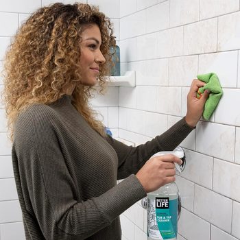 A woman cleans bathroom tiles with a green cloth, holding a spray cleaner in her other hand, surrounded by white tiled walls.