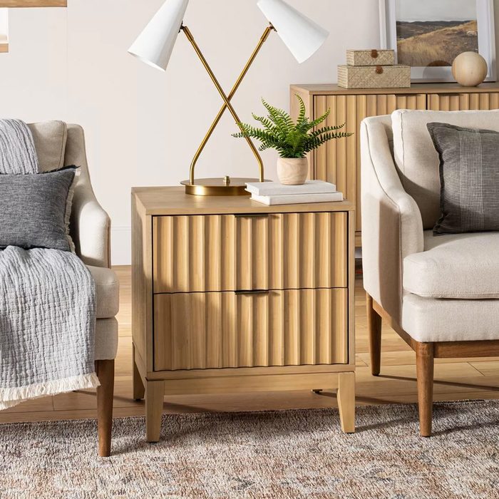 A wooden nightstand with ridged surfaces holds a lamp and a potted plant, situated between two cozy armchairs on a textured rug in a bright room.