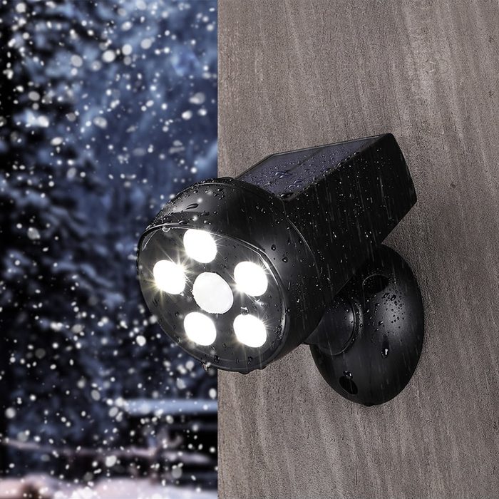 A black solar-powered light shines brightly against a wet, textured wall, surrounded by blurred snowflakes in the background, indicating a cold, outdoor winter scene.