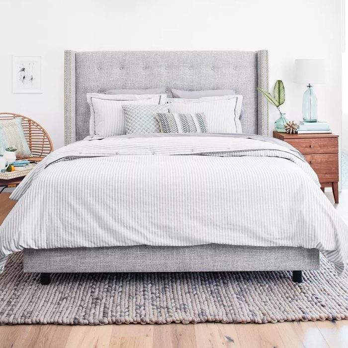 A gray-upholstered bed with striped bedding is centered, surrounded by a wooden nightstand, a rattan chair, and a light-colored rug on a wooden floor.