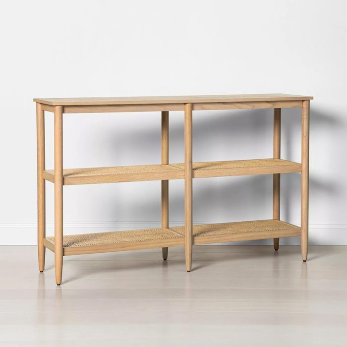 A wooden console table stands against a wall, featuring three shelves made of woven material, providing storage space in a minimalistic setting.