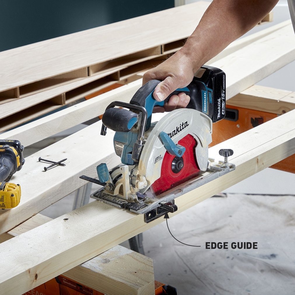 A circular saw is cutting through a wooden board while held by a person, surrounded by construction materials on a workbench with tools nearby.