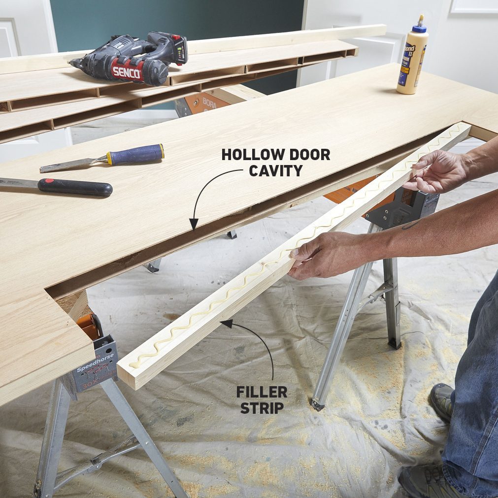 A person inserts a filler strip into a hollow door cavity, working on a wooden surface surrounded by tools and protective covering on the floor.