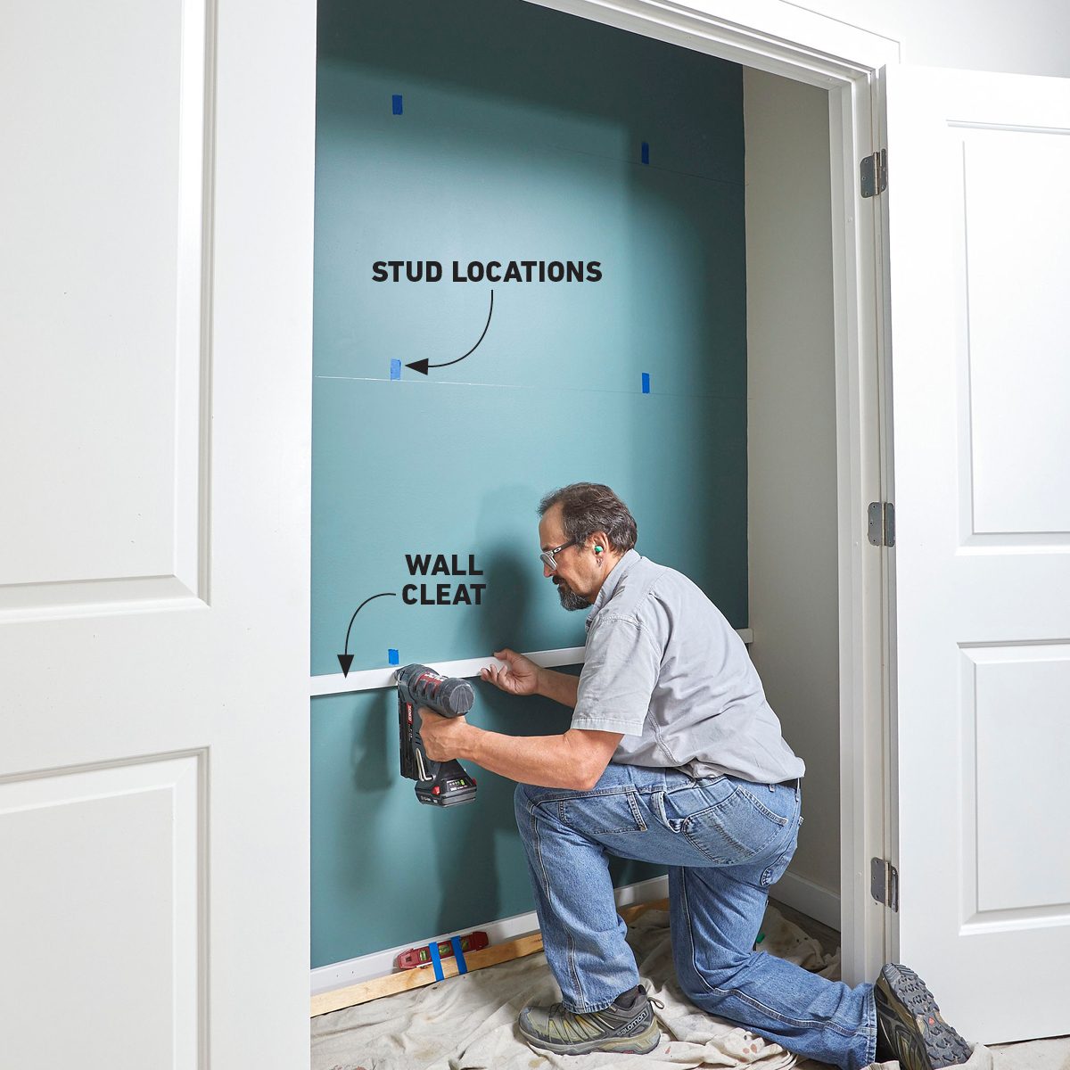 A man kneels, using a drill to attach a wall cleat on a blue wall, marked with stud locations, within a well-lit room.