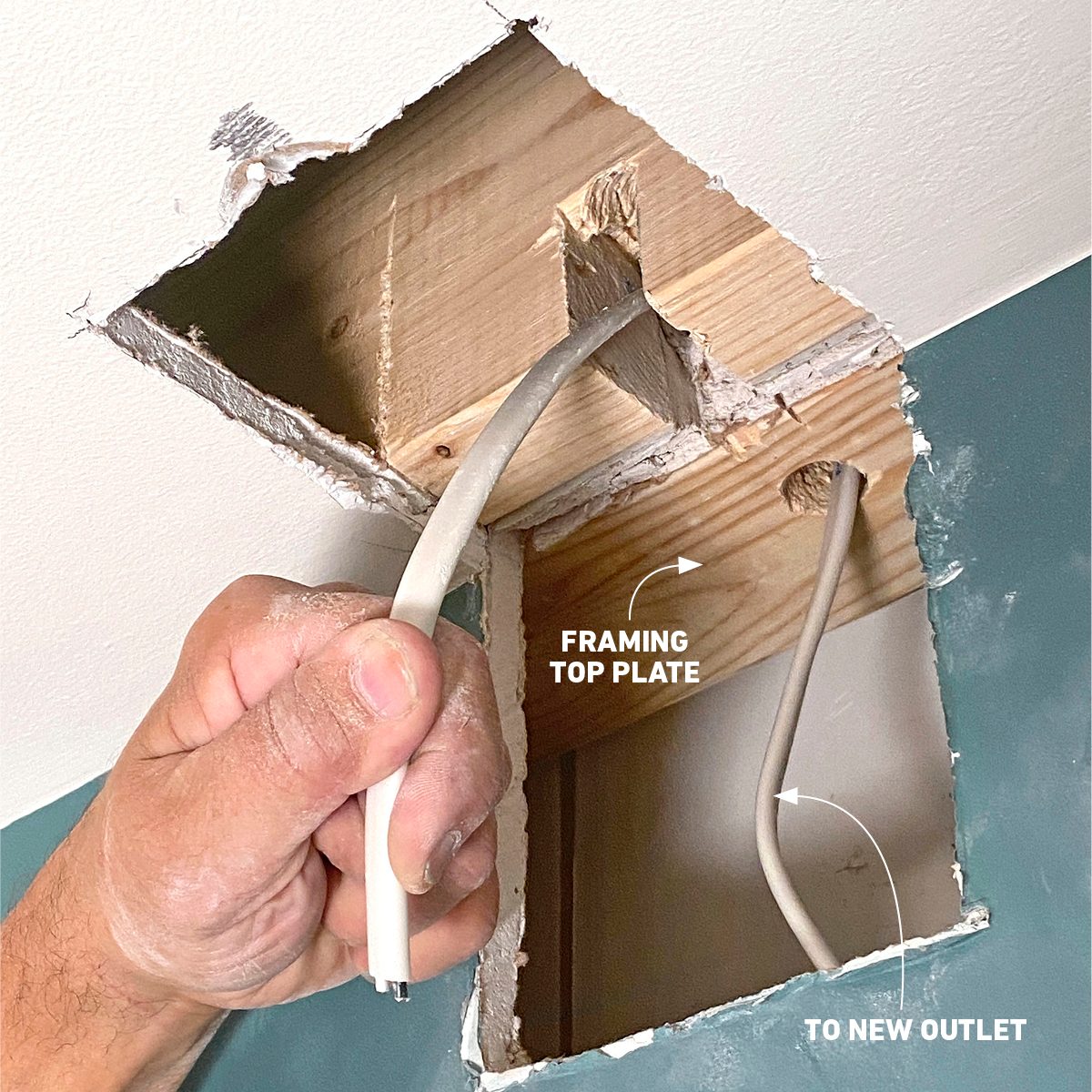 A hand holds electrical wire being routed through a cut-out section of a wall, near wooden framing towards a new outlet location.