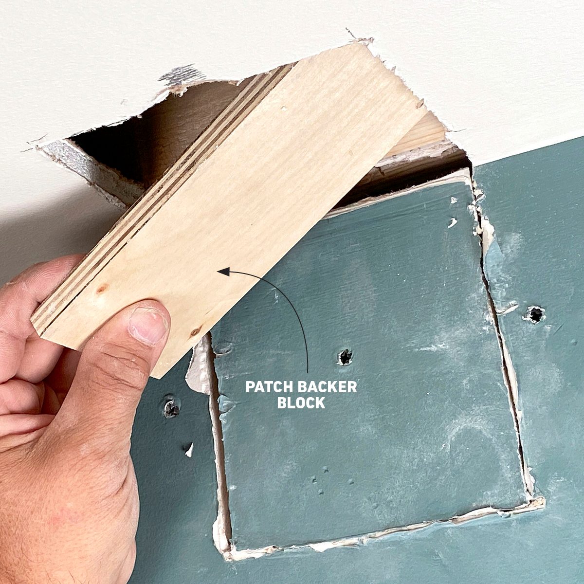 A hand holds a patch backer block against a wall, preparing to reinforce a cut-out area for repair in a partially damaged section.