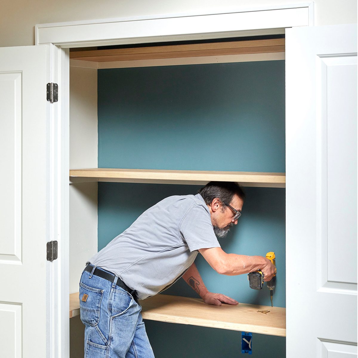 A man is using a drill to secure a shelf in a closet with blue walls, surrounded by white doors.