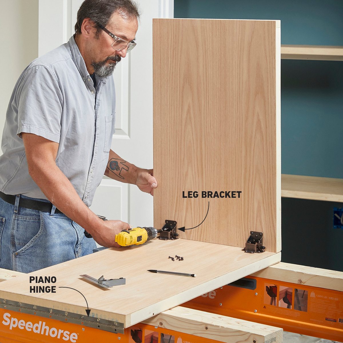 A man is using a power drill to attach a leg bracket and piano hinge to a wooden panel on a work surface in a room.