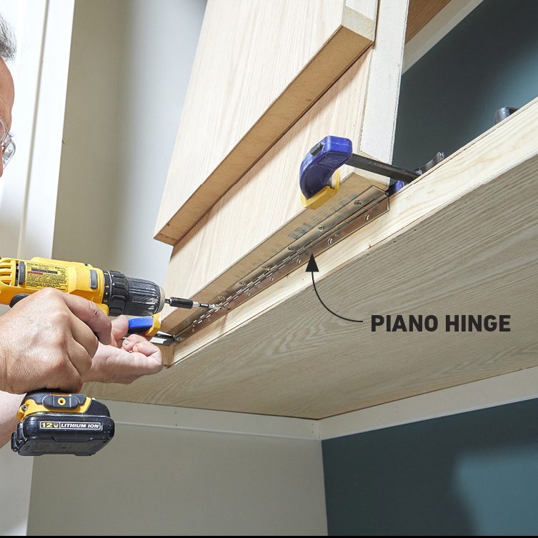 A hand holds a power drill, attaching a piano hinge to wood cabinets in a well-lit, indoor workspace. A clamp secures the hinge in place.
