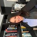 A person smiles and poses while leaning against a bookshelf filled with various books, creating a warm, welcoming atmosphere in a cozy space.