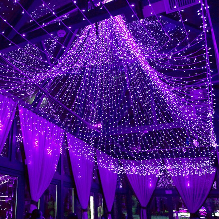 String lights hang from above, illuminating a space with purple hue. Soft fabric drapes frame the scene, enhancing the festive atmosphere.