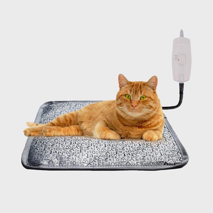 An orange cat lies comfortably on a patterned heating pad, which is plugged in and connected to a control unit for temperature adjustment.