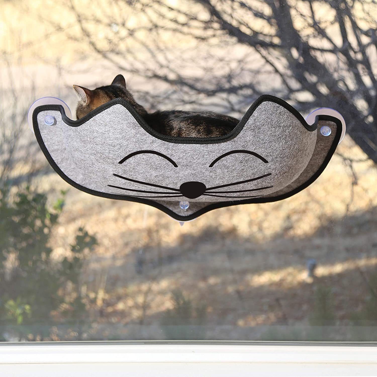 A cat rests in a gray felt hammock shaped like a smiling cat, secured to a window, with a blurred outdoor background of trees and grass.