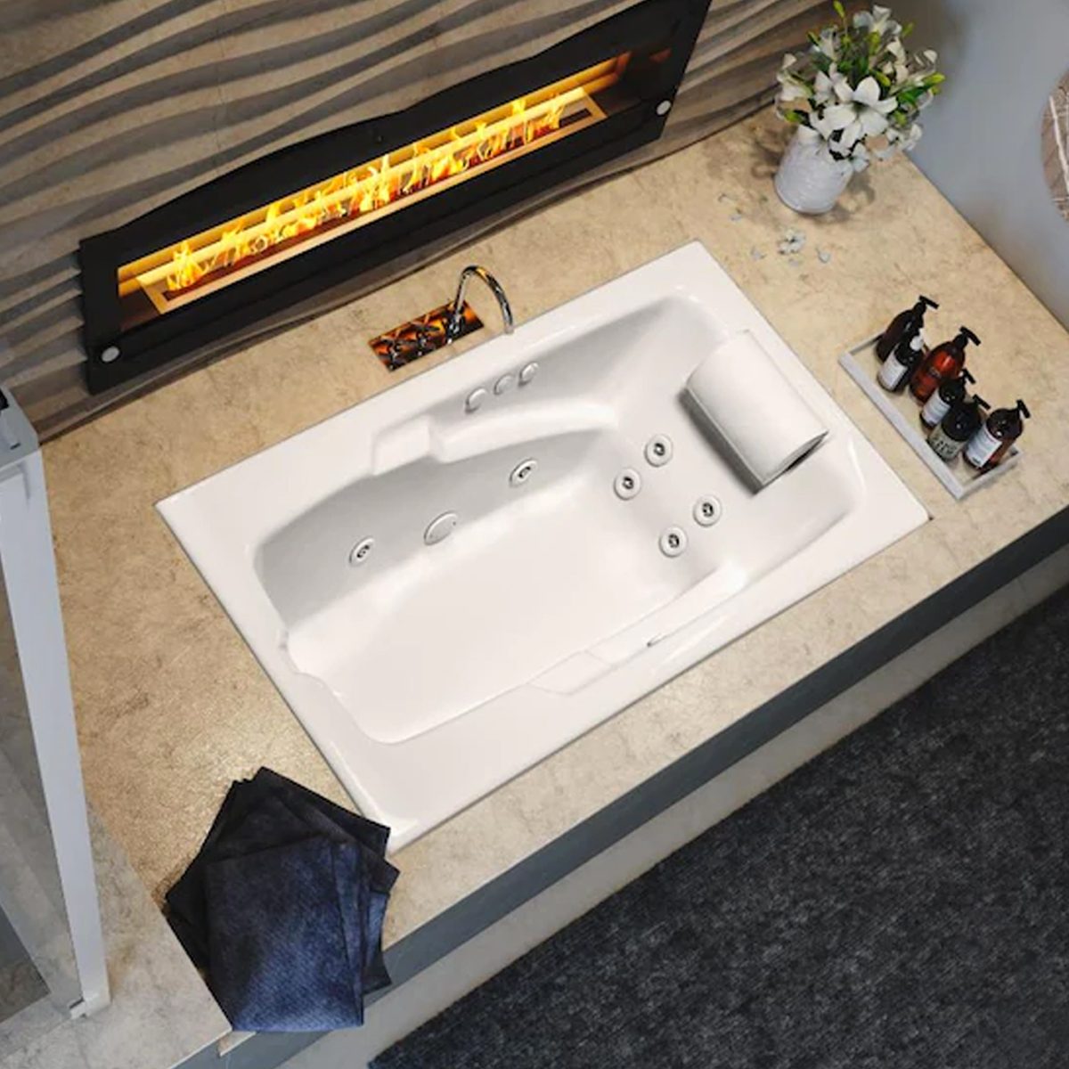 A white bathtub sits on a textured surface, with water jets visible. A fireplace glows above it, surrounded by decorative items and towels.