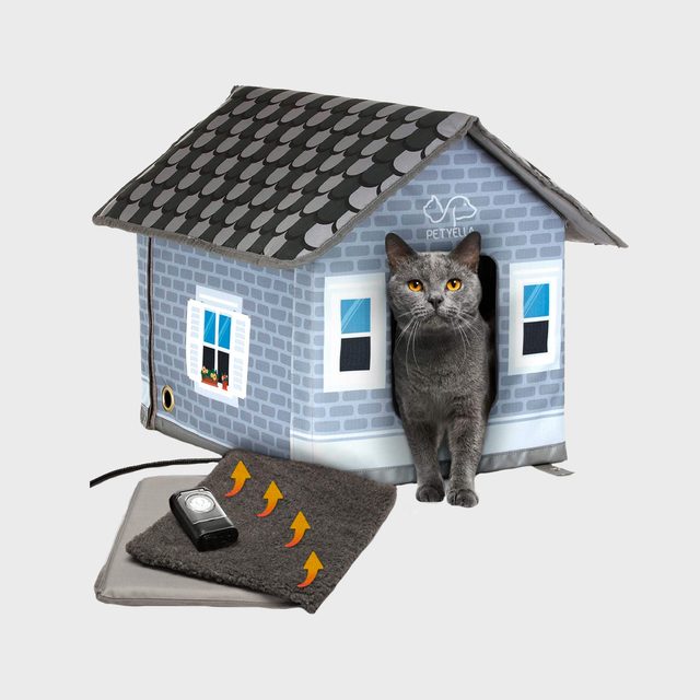 A gray cat sits in the entrance of a colorful, cartoonish house-shaped pet bed, alongside a heated mat with a device.