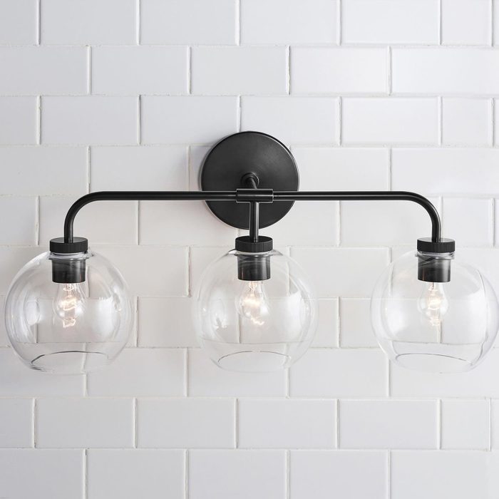A wall-mounted light fixture with three clear glass globes and exposed bulbs illuminates a clean, white tiled wall, adding a modern aesthetic to the space.