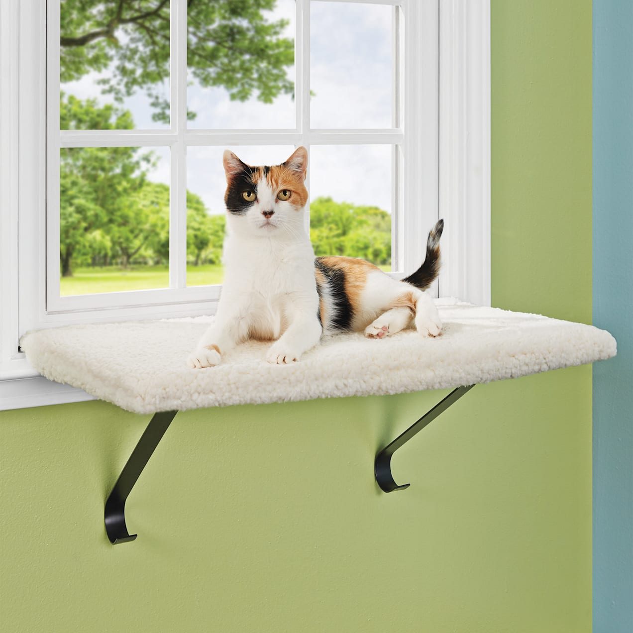 A calico cat lounges on a soft window perch, gazing out at a lush green landscape through a large window.