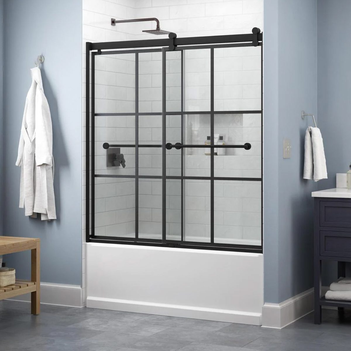 A glass shower enclosure with black frame slides open. A light blue wall surrounds it, complemented by a wooden bench and white towels nearby.