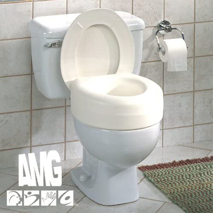 A white toilet with a raised seat is positioned on tiled flooring, accompanied by a toilet paper roll on a nearby holder and a textured mat beside it.