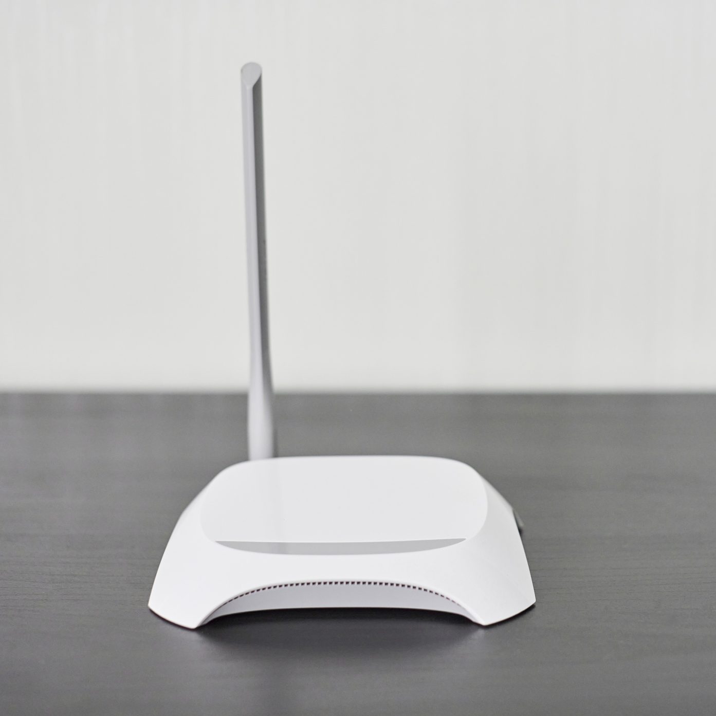A white router stands upright on a dark surface, with a long antenna extending upwards against a light, neutral background.