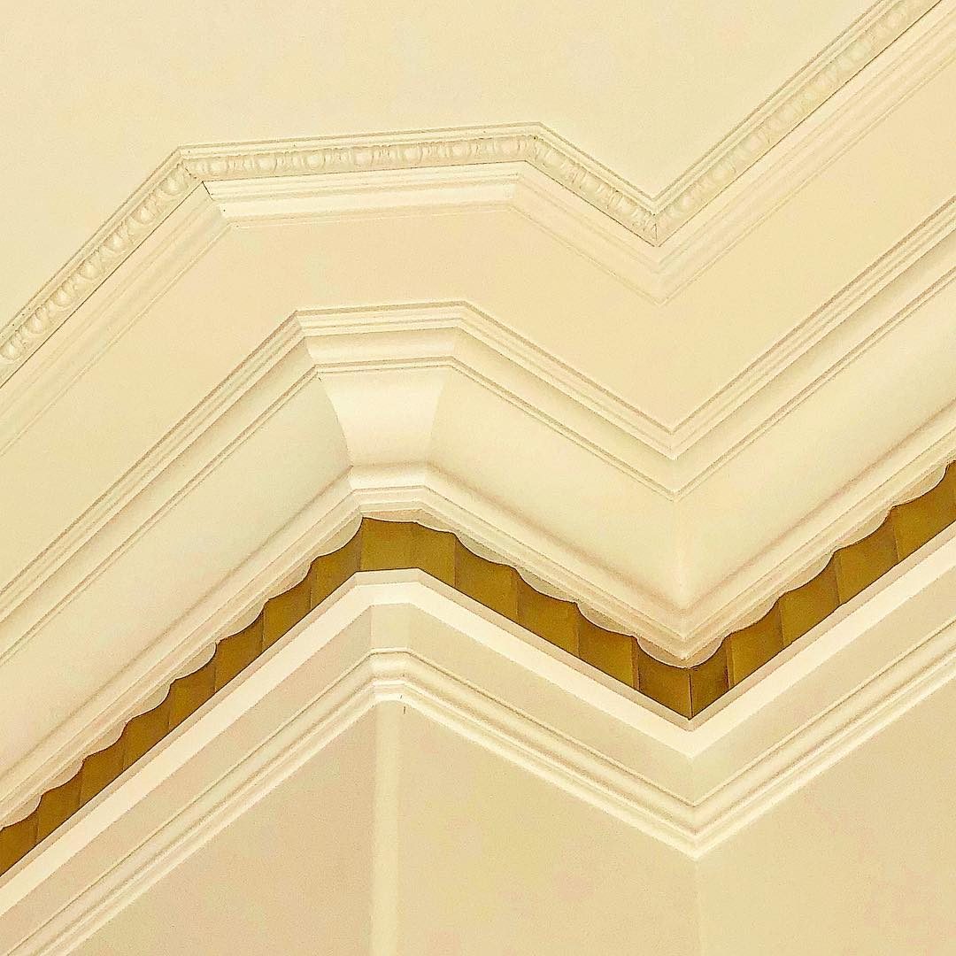 A decorative ceiling corner features layered molding, showcasing intricate detailing and a scalloped edge, all set against a light, neutral background.