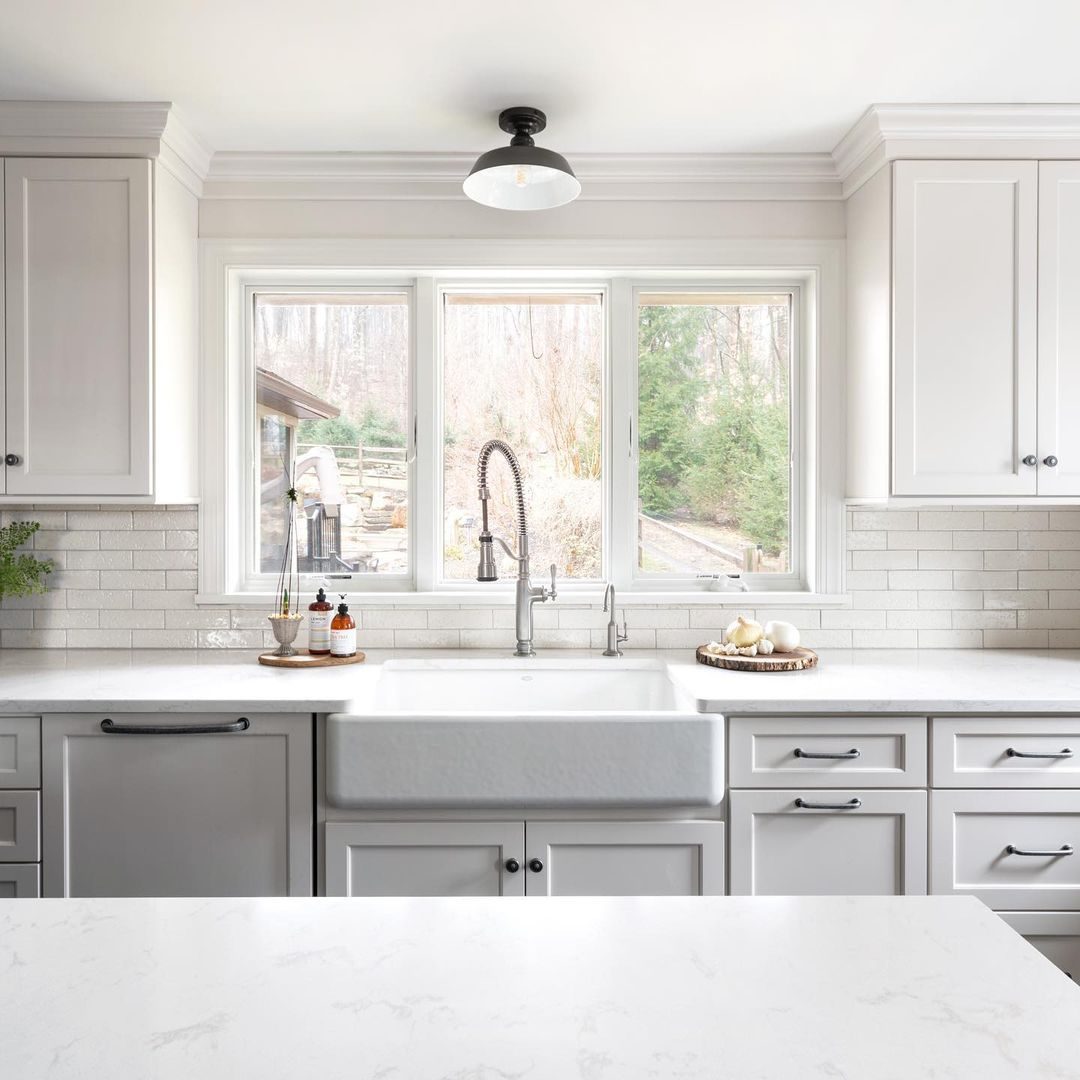 A farmhouse sink sits beneath large windows, while a stylish faucet and decorative items enhance a bright, modern kitchen with light cabinetry.