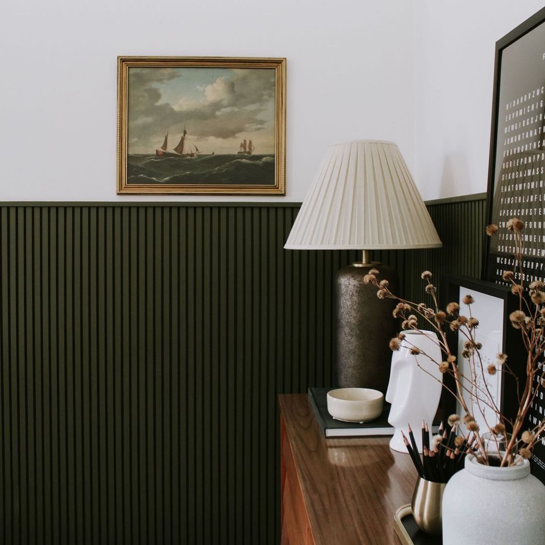 A lamp stands on a wooden dresser, accompanied by decorative items. A painting of sailing ships hangs on a green paneled wall in a bright room.