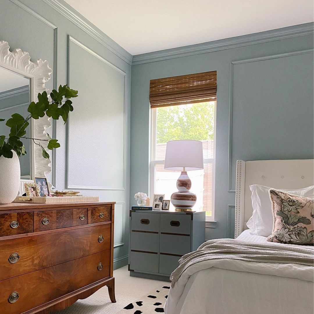 A wooden dresser holds decorative items; a lamp illuminates a nightstand beside a bed, with a window revealing greenery outside in a light blue room.