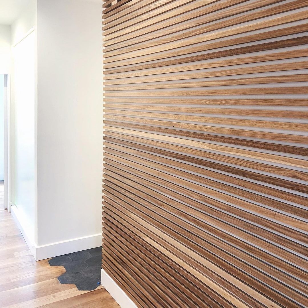 A wooden slatted wall stands upright, featuring alternating light and dark stripes. A smooth floor leads to a bright, open room beyond.