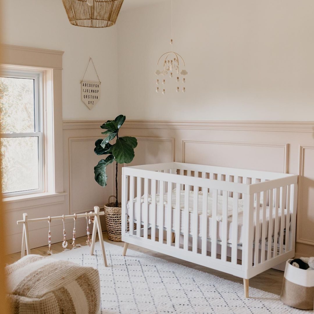 A white crib rests in a softly lit nursery, surrounded by a plant and a play gym, on a patterned rug under a natural light source.