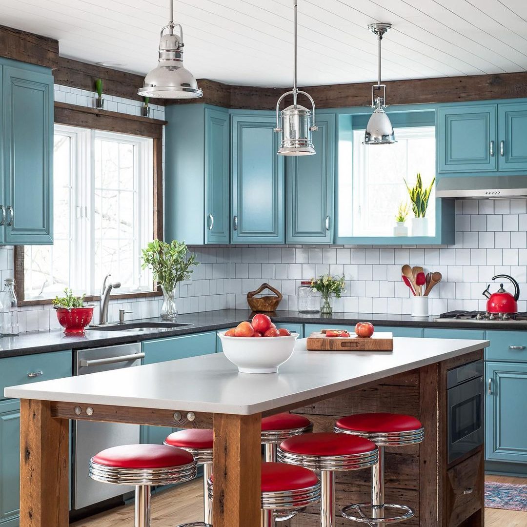 A modern kitchen features blue cabinetry and a white countertop. A bowl of apples sits on the island, surrounded by red bar stools.