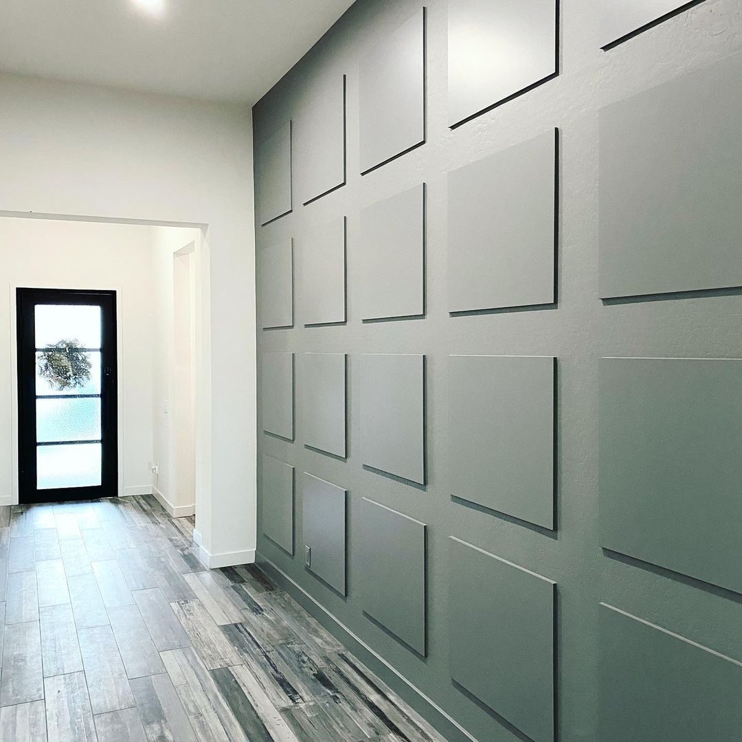 A gray wall features evenly spaced rectangular panels. Light-colored flooring extends towards a doorway with a glass door, leading outside to a well-lit area.